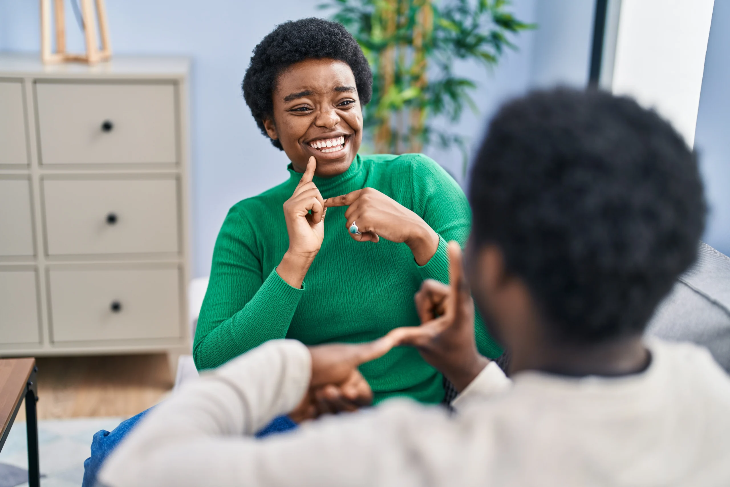 Woman in green turtleneck laughing while signing to a man sitting across from her.
