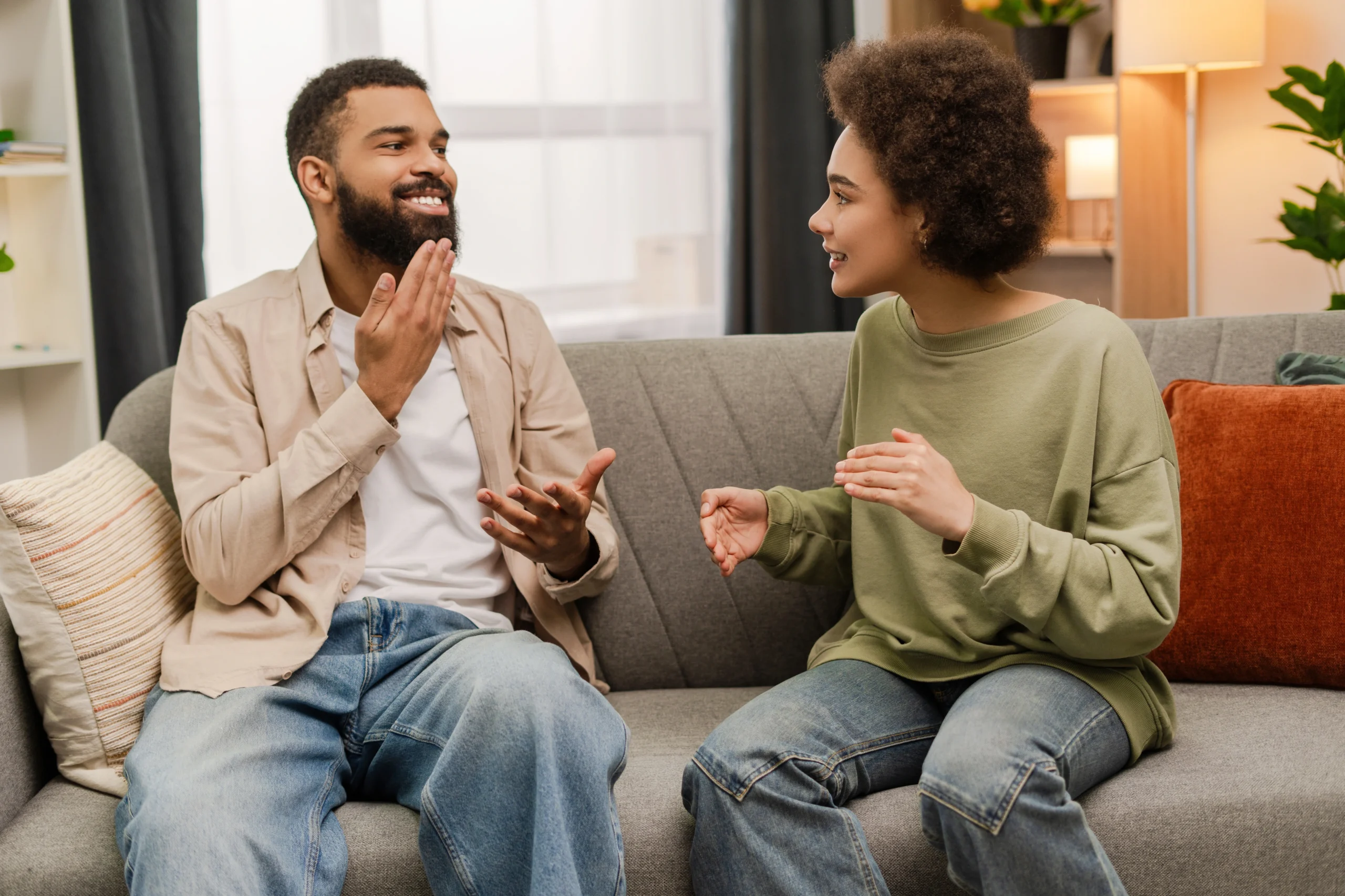 Man on a couch in a tan button down shirt using sign language to communicate with the woman sitting next to him